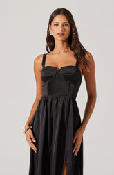 A woman is wearing a sleeveless black dress with wide shoulder straps. The bodice features vertical ribbed fabric panels and built-in, rounded cup-like design elements at the bust. The waistline gathers into a flowing skirt with a slit on one side.